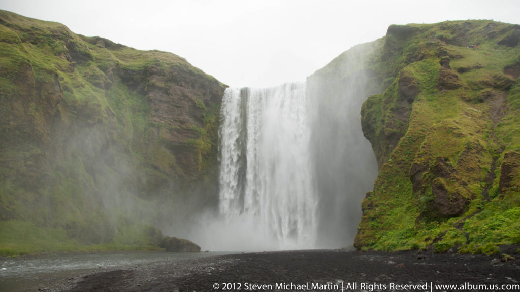 A wide waterfall plunges between green cliffs into a dark river channel.