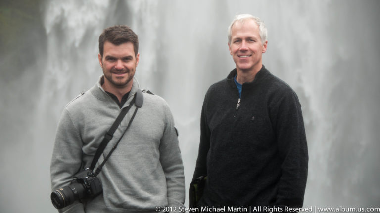 Two men pose together outdoors, one wearing a gray sweater with a camera and the other a dark jacket.