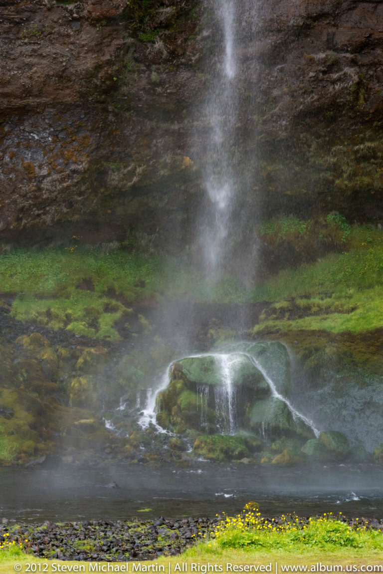 A narrow waterfall spills over a rounded rock shelf into a shallow pool beneath mossy cliffs.