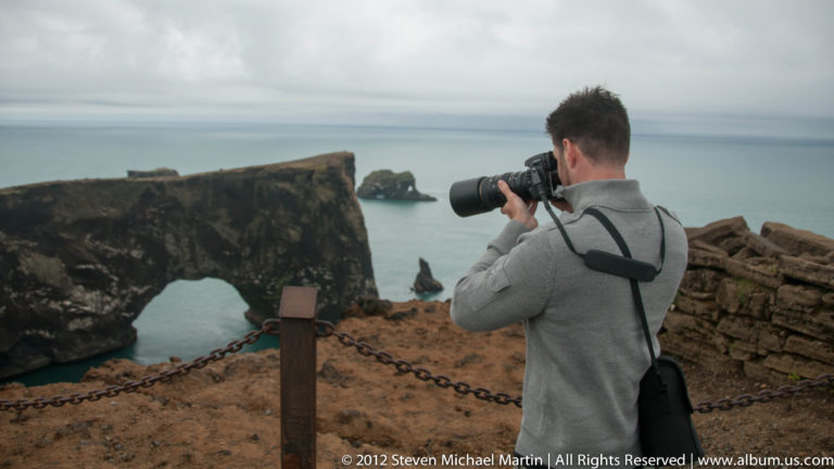 A photographer stands at Dyrhólaey beside the large basalt sea arch, aiming toward offshore rock stacks.