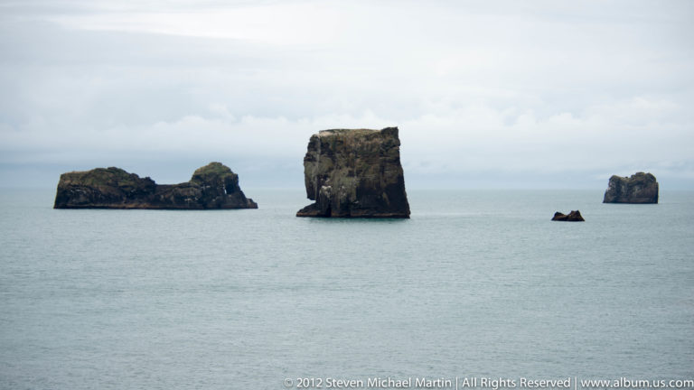 Three dark rock stacks rise from calm water beneath a pale, level horizon.