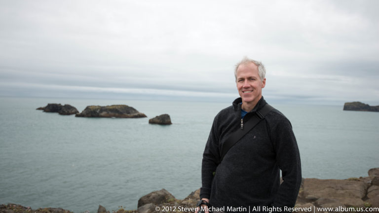 A man in a dark jacket stands on a rocky shore with low sea stacks behind him.