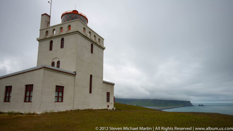 Dyrhólaey lighthouse, a pale square tower with red trim, stands on the grassy coastal headland.