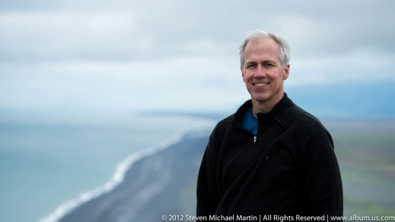 A smiling man in a dark jacket stands before a pale sea dotted with low rocks.