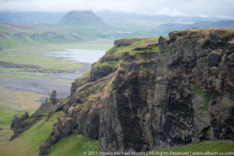 A fractured basalt cliff overlooks a broad green valley, distant water, and hazy mountains.