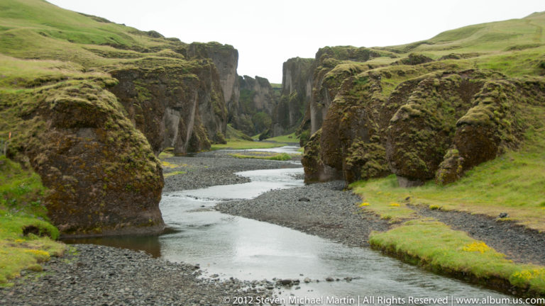 A shallow river winds between tall dark canyon walls topped with green grass.
