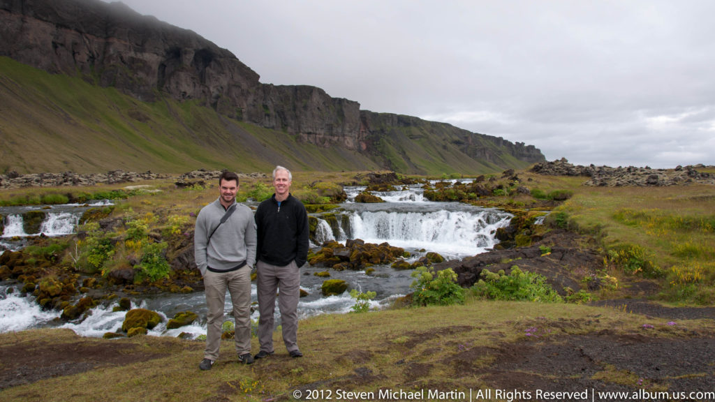 Two men stand beside whitewater below a long green-topped canyon wall.