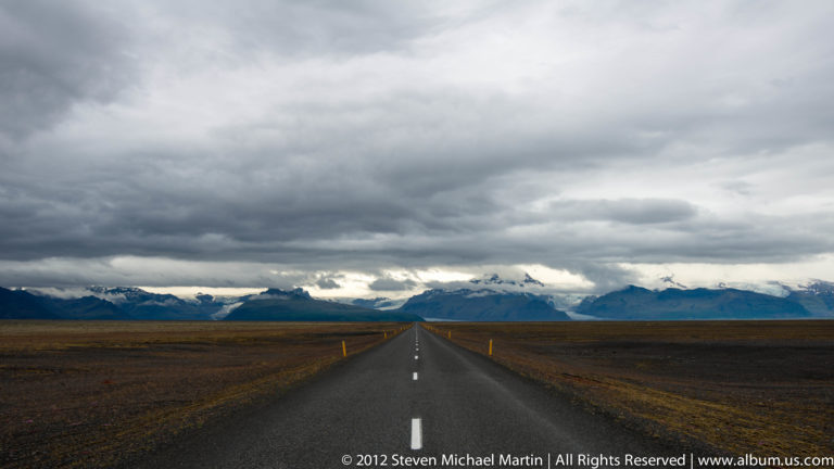 A straight paved road crosses a dark plain toward blue mountains beneath low storm clouds.