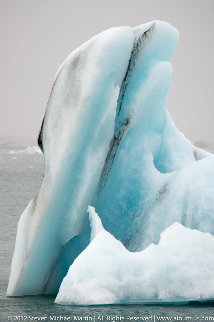 Curved white-and-blue iceberg rising from Jökulsárlón glacier lagoon behind smaller pieces of floating ice.