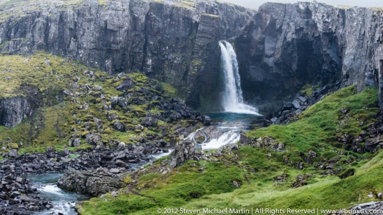 A narrow waterfall falls through a rocky fissure into a dark pool and lower cascade.
