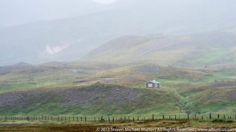 A small red house stands in a broad grassy valley with hazy hills and a fence in the foreground.