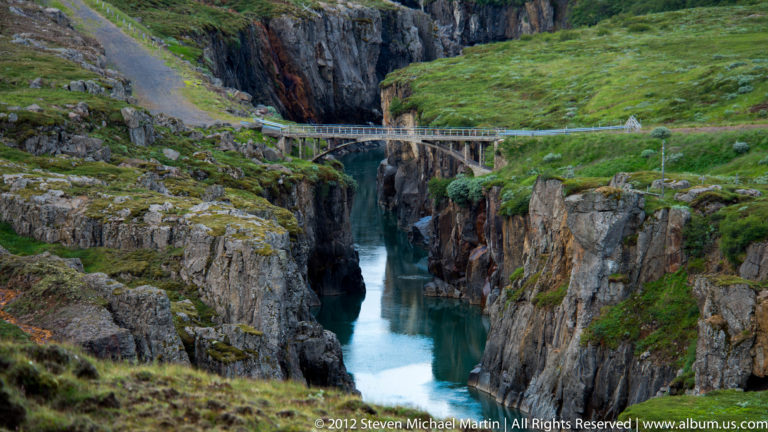 A footbridge crosses a turquoise river cutting through a steep, grass-topped rock gorge.