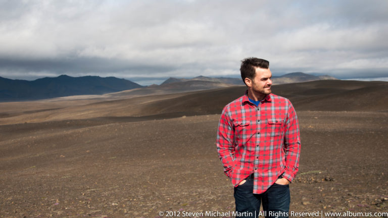A man in a red plaid shirt stands on dark gravel with low mountains beneath an overcast sky.