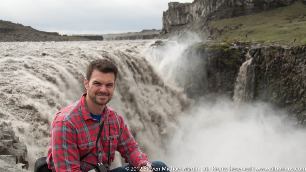 A seated man in a red plaid shirt poses beside a powerful waterfall and dark cliff.