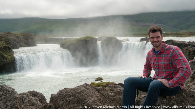 A traveler sits on rocks before the broad horseshoe-shaped cascade of Goðafoss.