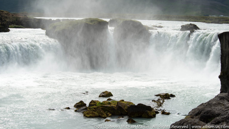 Close view of Goðafoss pouring over dark basalt ledges into a misty basin.
