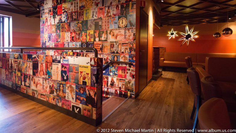 Colorful Playboy covers and posters cover a wall beside a dim lounge with tables and pendant lights.