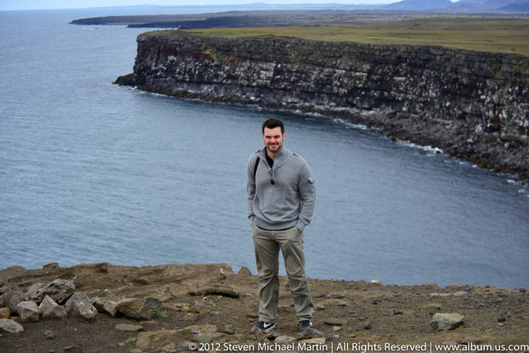 A man in a gray pullover stands on a rocky headland above calm water and a distant cliff.