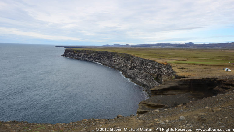 A long dark coastal cliff with a grassy top curves above a narrow rocky shore.