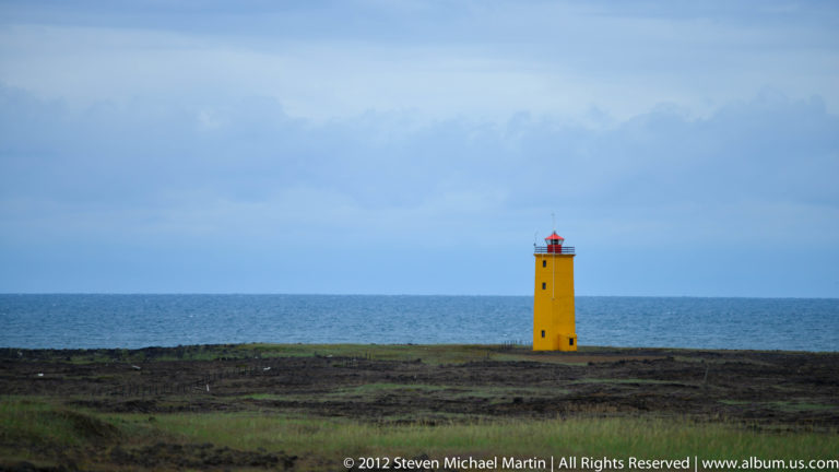 A yellow lighthouse with a red lantern stands on a flat dark shore beside the sea.
