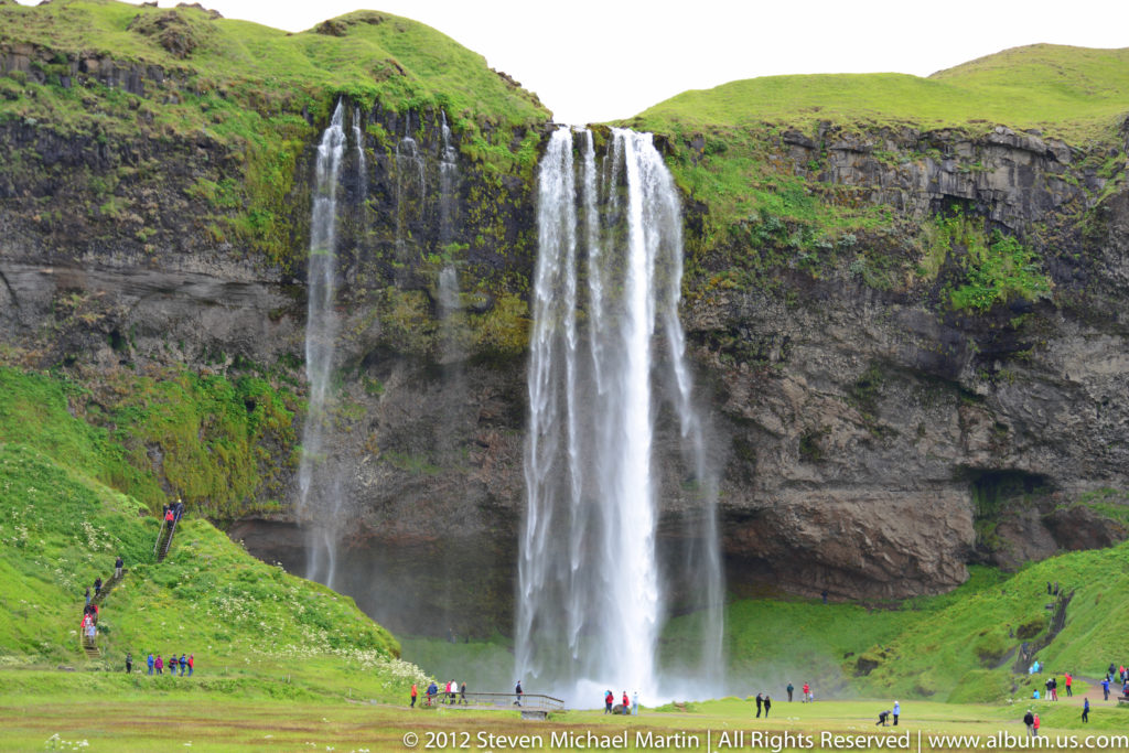 Several waterfalls descend over a tall cliff beneath a grassy plateau, with small visitors below.