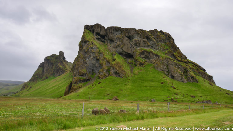 A steep dark rock mound with green slopes rises from a flat fenced pasture.