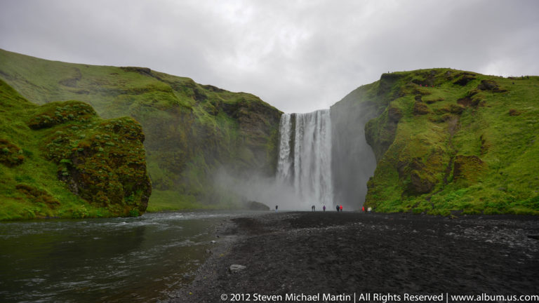 A broad curved waterfall divides around dark rocks and spreads into a pale pool.