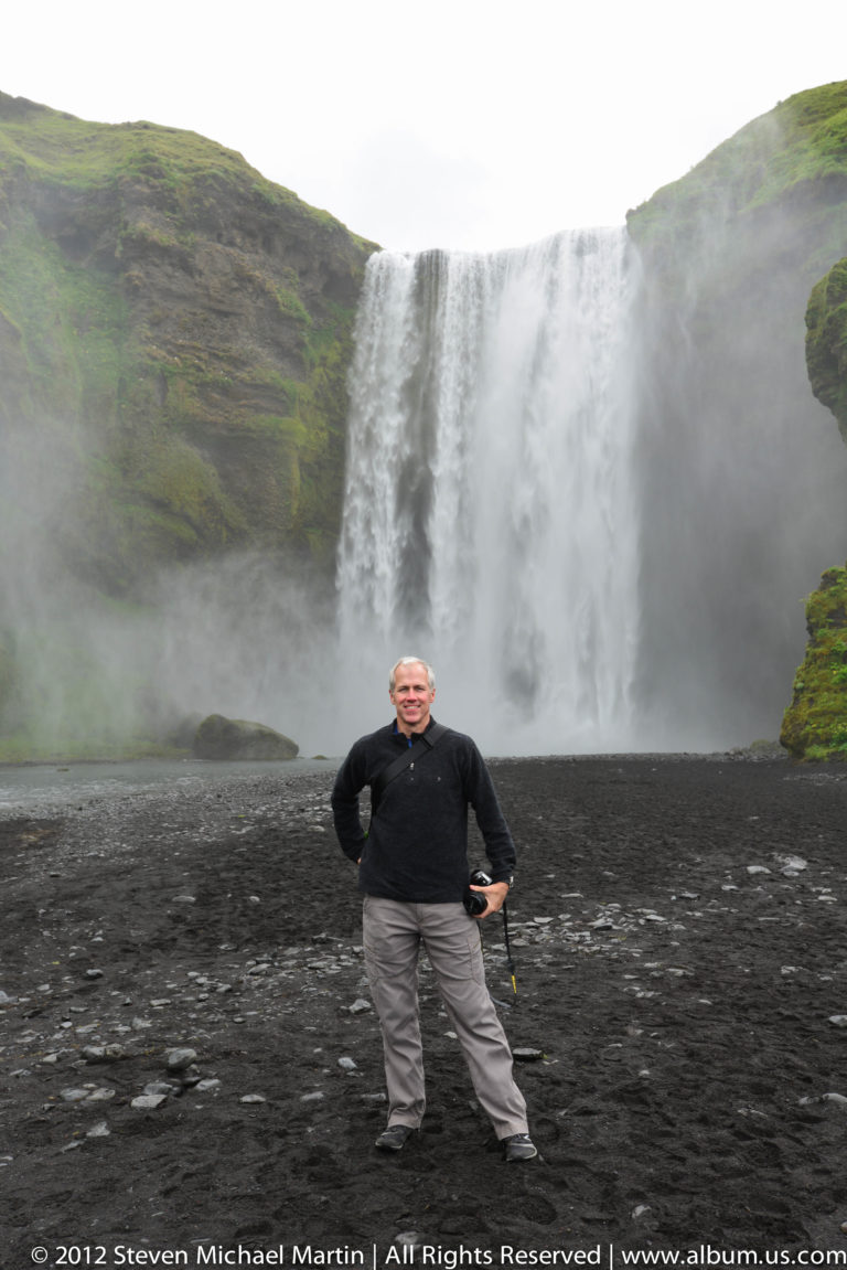 A traveler stands on black gravel before the broad white curtain of Skógafoss.