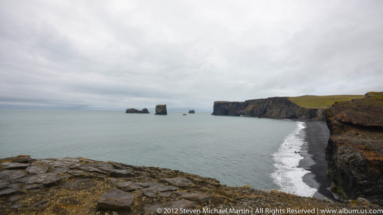 A dark coastal cliff and black beach curve toward several offshore stacks under a cloudy sky.