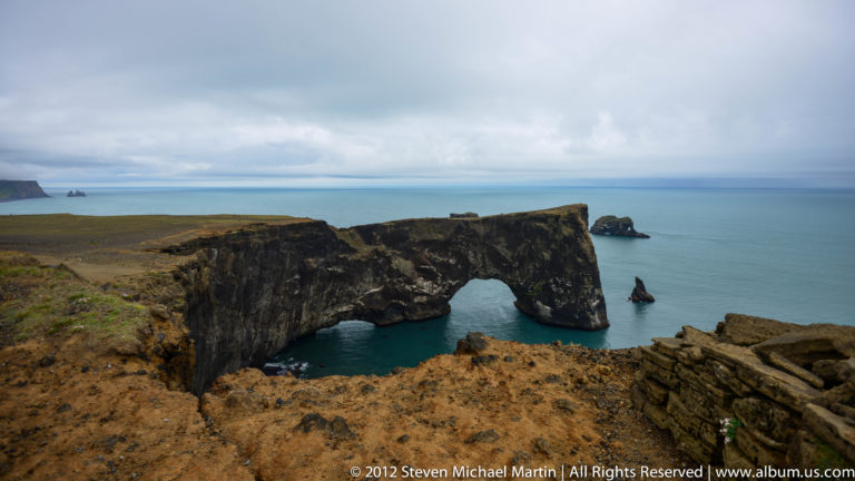 Wide view of Dyrhólaey’s sea arch beneath a flat-topped basalt headland with an offshore stack.