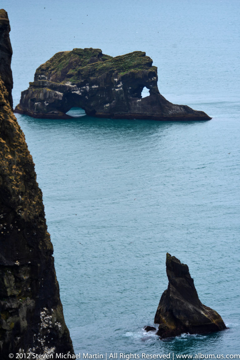 An angular sea stack rises from blue water beside a dark cliff edge.