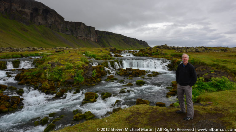 A man stands beside whitewater flowing around dark boulders below a long grassy cliff.