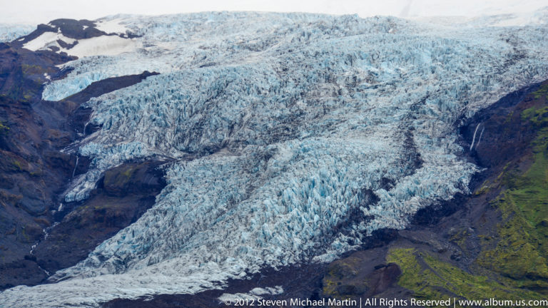 Close view of a glacier’s rough blue-white surface cut by dark crevasses and debris bands.