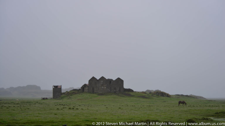 Roofless stone ruins stand on a low rise with a horse grazing in the foreground pasture.