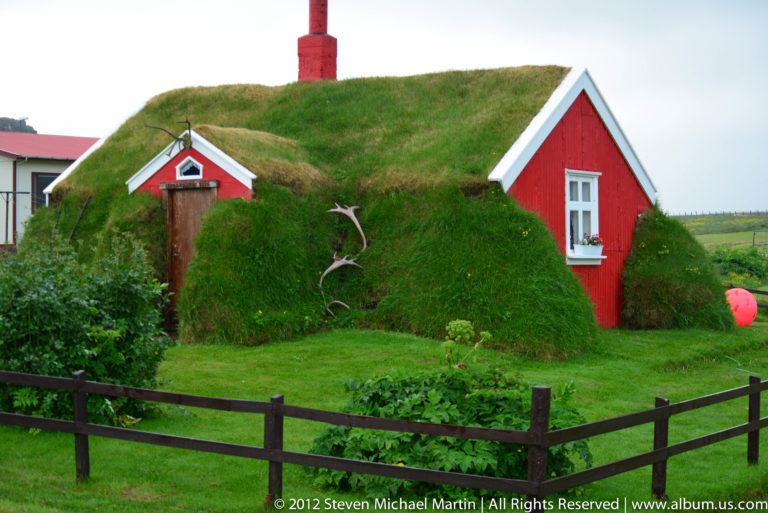 Red timber buildings with white trim sit beside a steep grassy bank and dark foreground fence.