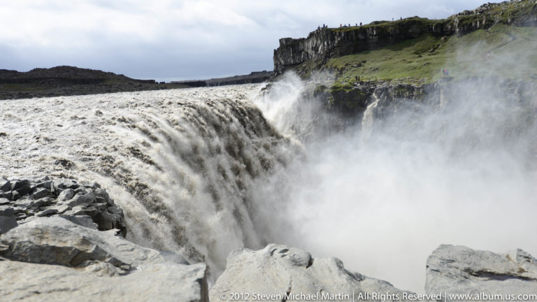 Visitors stand near the grassy rim beside Goðafoss and its basalt gorge.