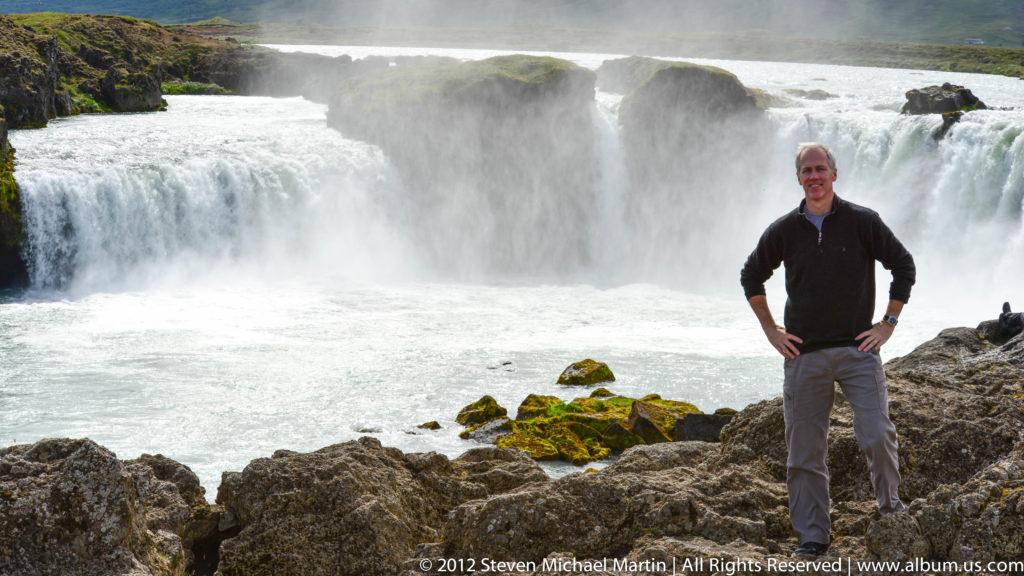 A traveler stands on a rocky riverbank before the broad cascade of Goðafoss.