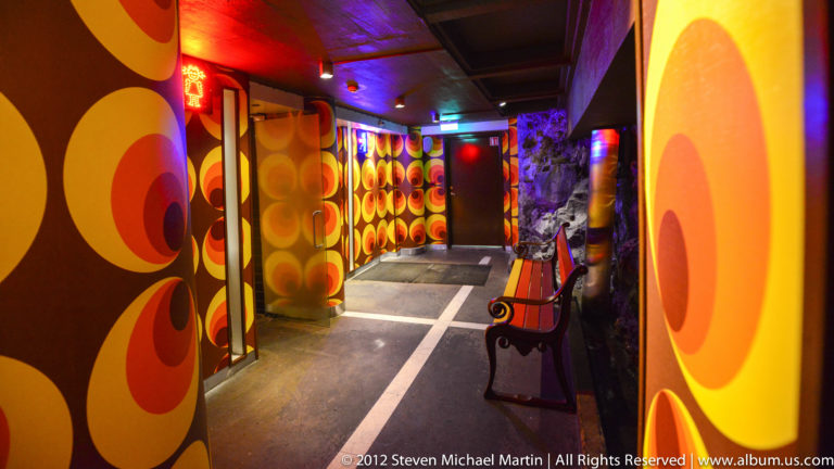 An empty corridor with large orange wall motifs, colored ceiling lights, and a chair beside the passage.