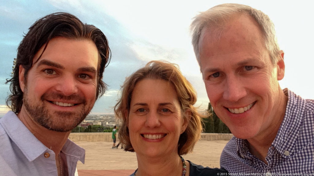 Three smiling adults take a close selfie with Barcelona faintly visible behind them.