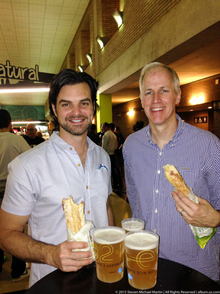 Two men hold sandwiches and stand behind several beers in a crowded food venue.