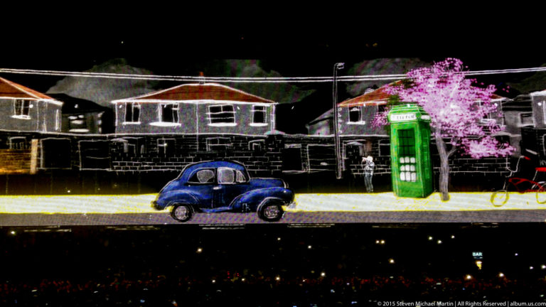Concert stage projection of houses, a blue car, green kiosk, bicycle, and bright tree.