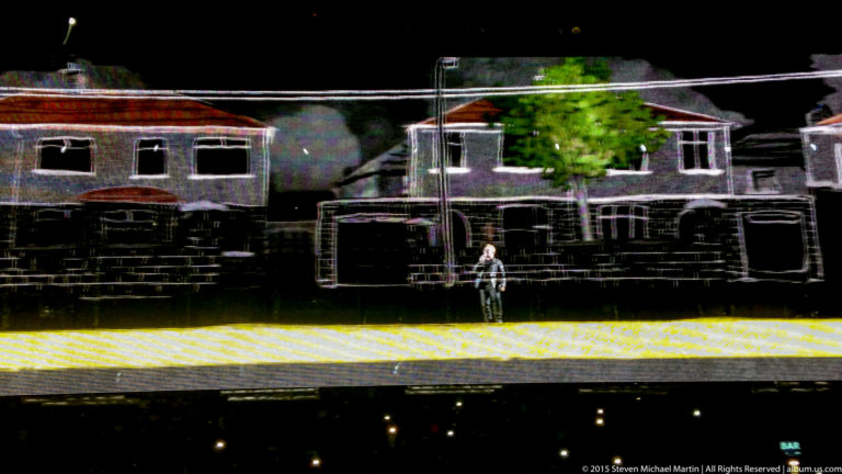 Wide concert stage projection of outlined houses with a small standing figure at center.