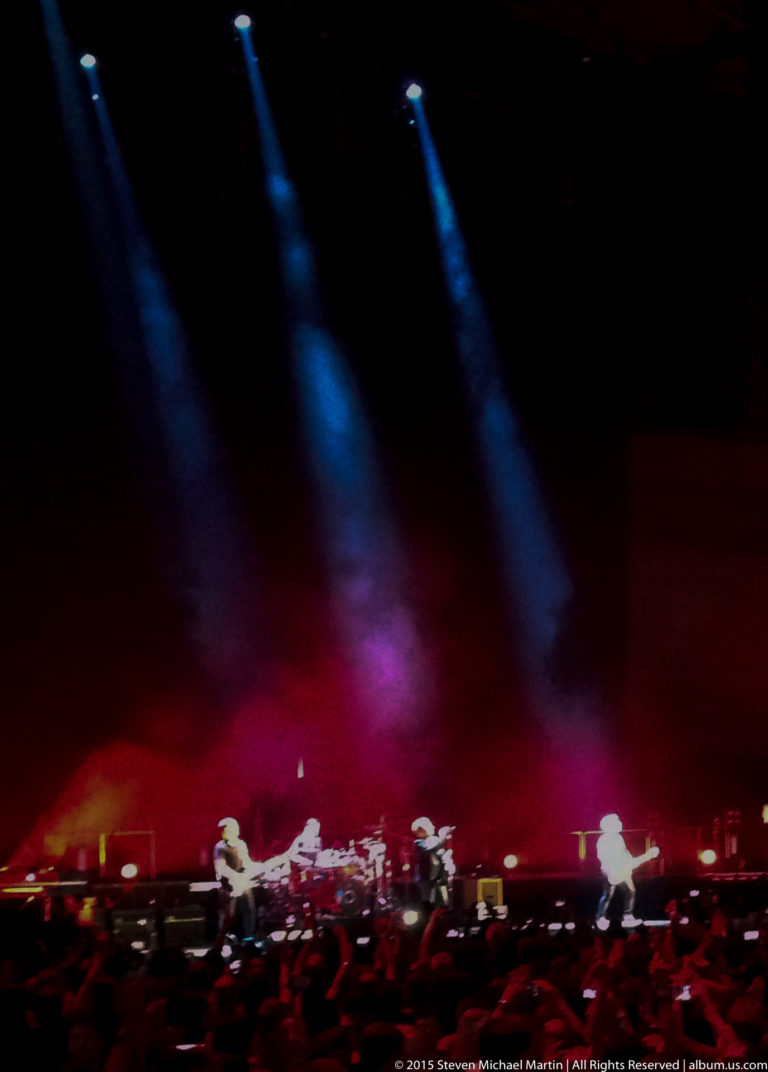 Wide concert stage with small performers beneath red light and three blue spotlights.