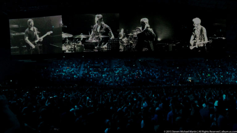 Black-and-white performer screens above a dense concert crowd illuminated in blue.