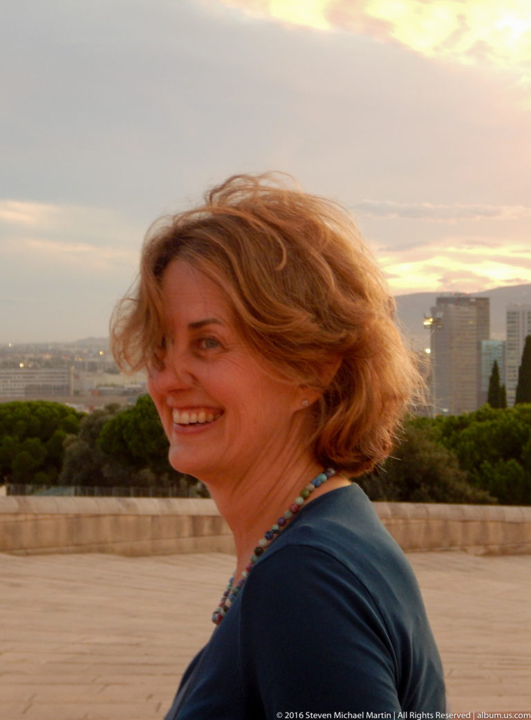 Smiling woman in profile at sunset with a softly focused Barcelona skyline behind her.