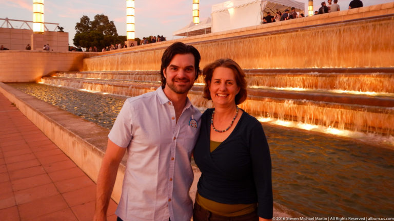 Man and woman pose beside a long amber-lit cascading fountain on Montjuïc at dusk.