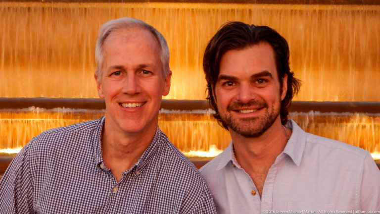 Two smiling men pose closely in front of an amber-lit cascading fountain on Montjuïc.