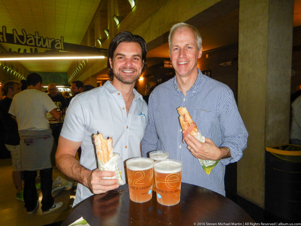 Two men hold sandwiches beside several beers at a crowded indoor table.