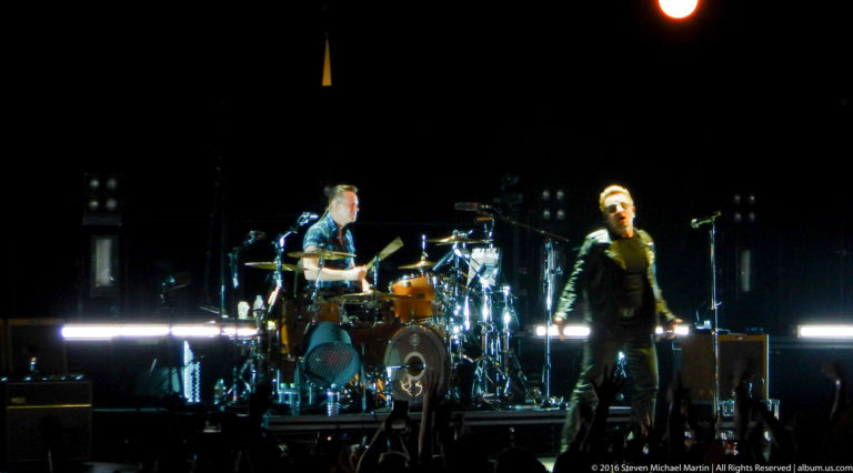 Male singer and drummer perform on a dark arena stage under bright white lights.