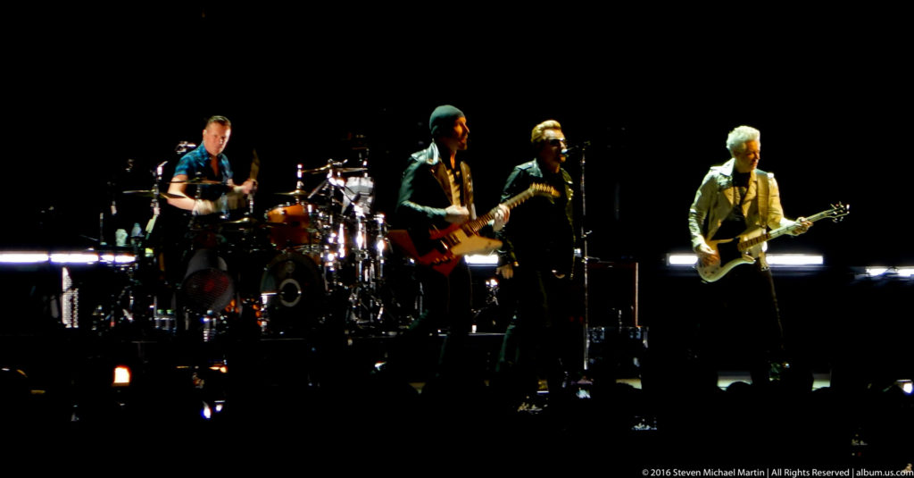 Four-piece rock band performs on a dark stage with drums and guitars under spotlights.